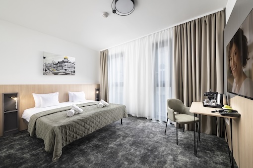 Tágas Deluxe hálószoba franciaággyal, ablak melletti ülősarokkal és szürke dekorációval.